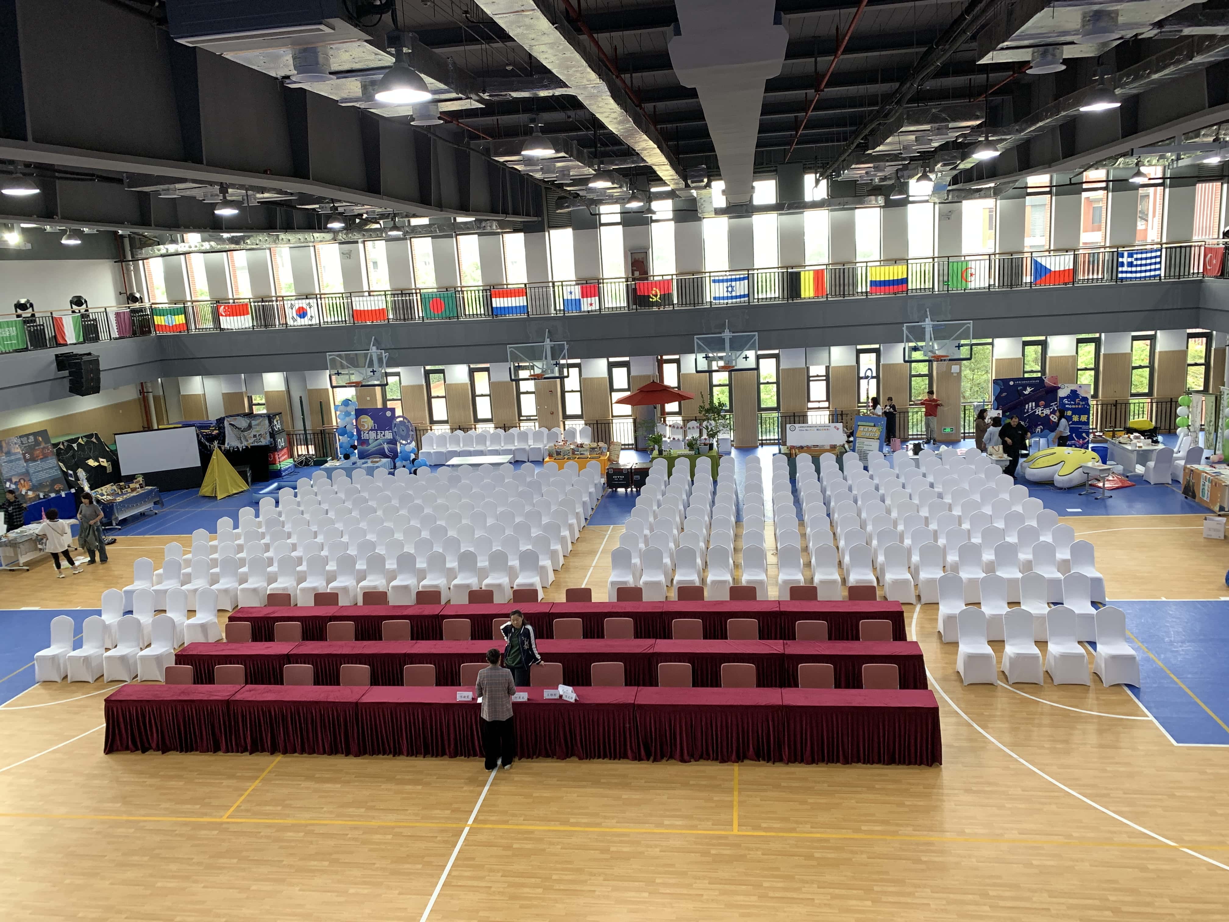 学校体育馆大型活动 — 白色椅套椅配深红色桌布长桌，各国国旗装饰国际学校活动